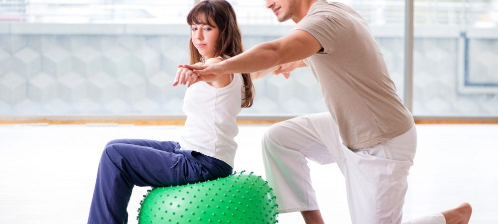 A girl sitting on a medicine ball next to her physical therapist with her arms pointing out.