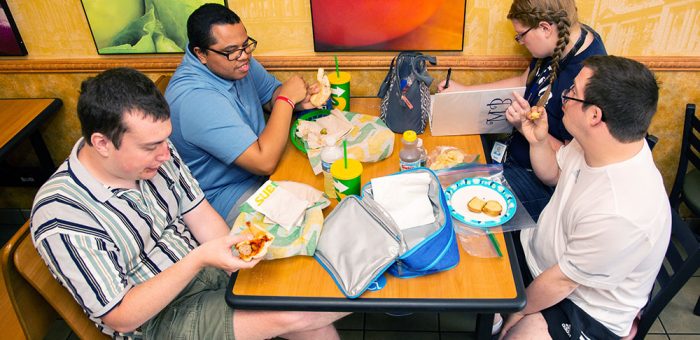 Four young adults are seen sitting in a sandwich shop, eating a meal together and talking.