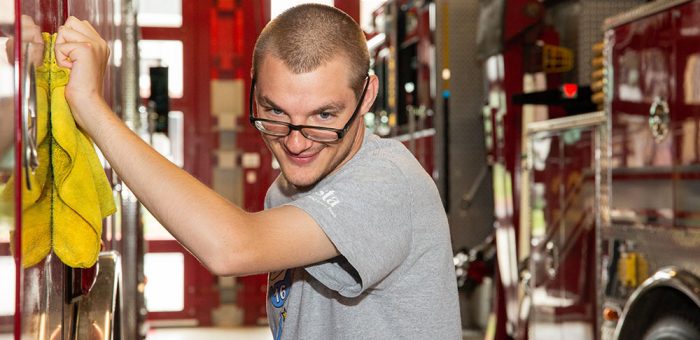 A young man is seen polishing a fire truck in a fire station. He is looking at the camera happily.