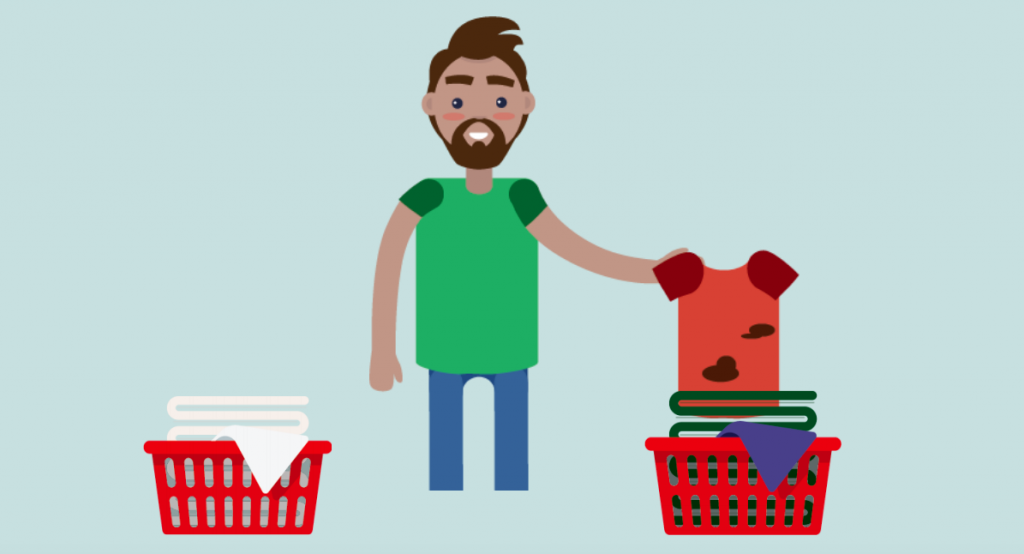 A man stands in the middle of two laundry baskets, holding a dirty shirt over the basket on the right.